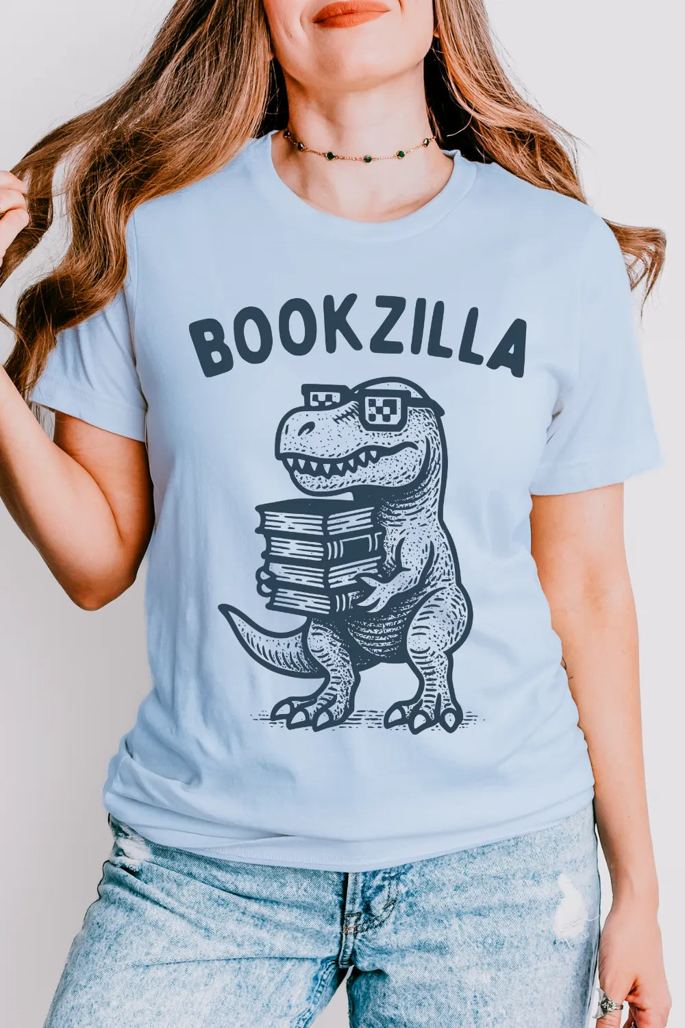 Person wearing a funny bookish t-shirt in light blue with 'BOOKZILLA' text and a dinosaur graphic holding books.