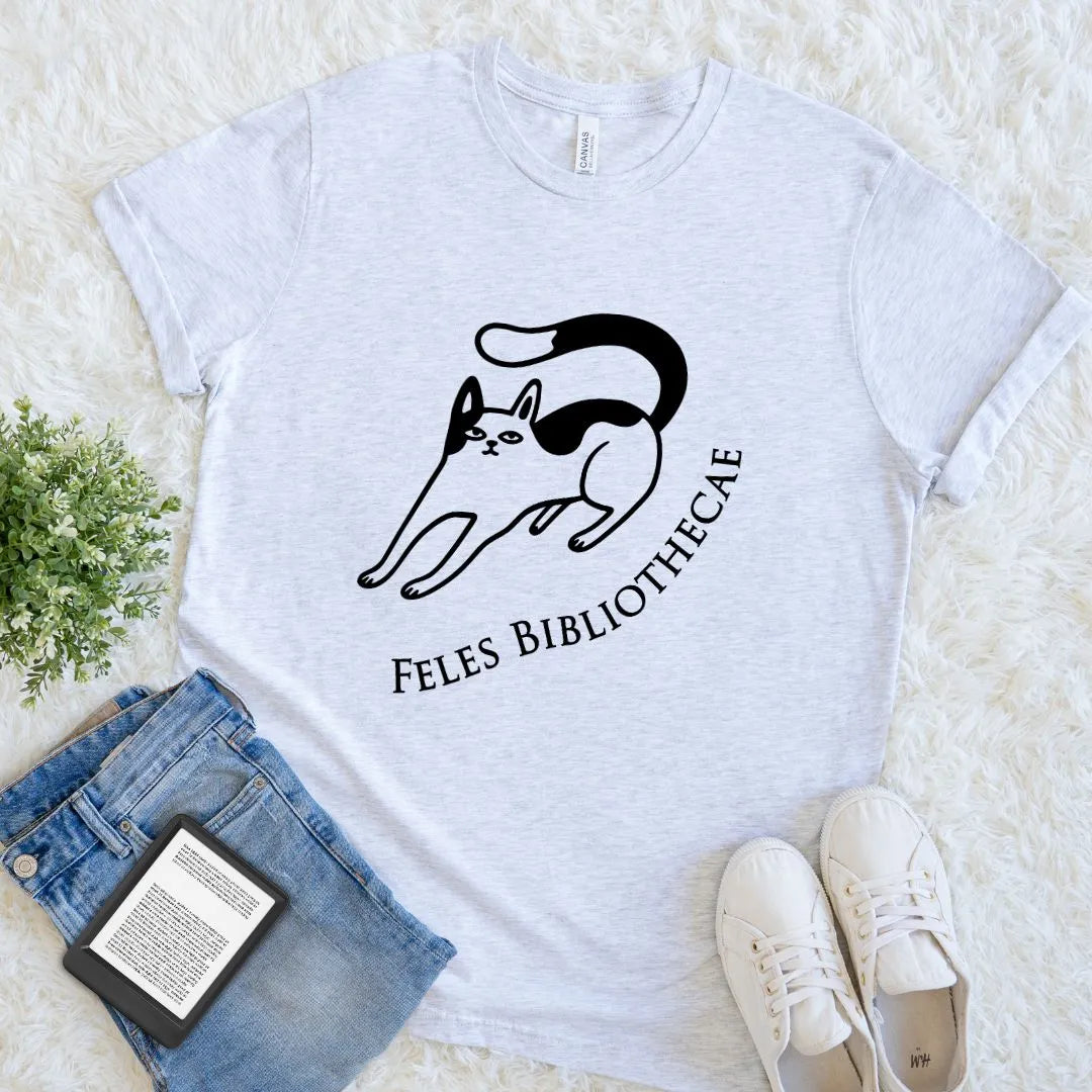 A light heather gray t-shirt with a black graphic of a cat and the text 'FELES BIBLIOTHECAE' underneath, styled in a flat lay on a white faux fur background with blue jeans, a book, and white sneakers.