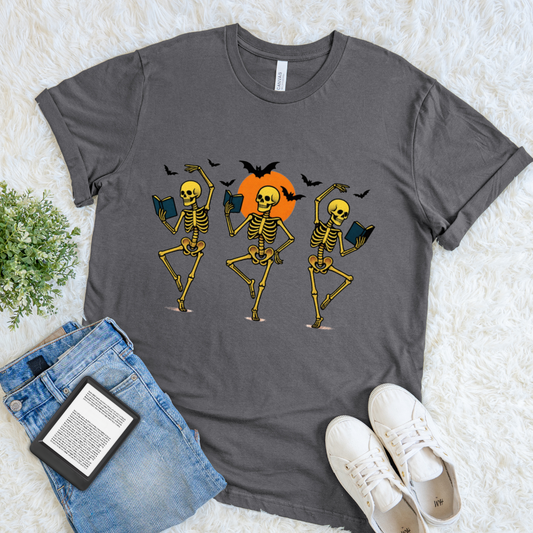 A dark gray halloween bookish graphic t-shirt featuring three dancing skeletons with books and bats on an orange sun, styled in a flat lay with blue jeans, a book, and white sneakers on a white faux fur background.