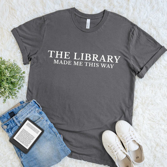A dark gray graphic t-shirt for readers with The Library Made Me This Way quote in bold cream serif typography, styled in a flat lay with blue jeans, a book, and white sneakers on a white faux fur background.