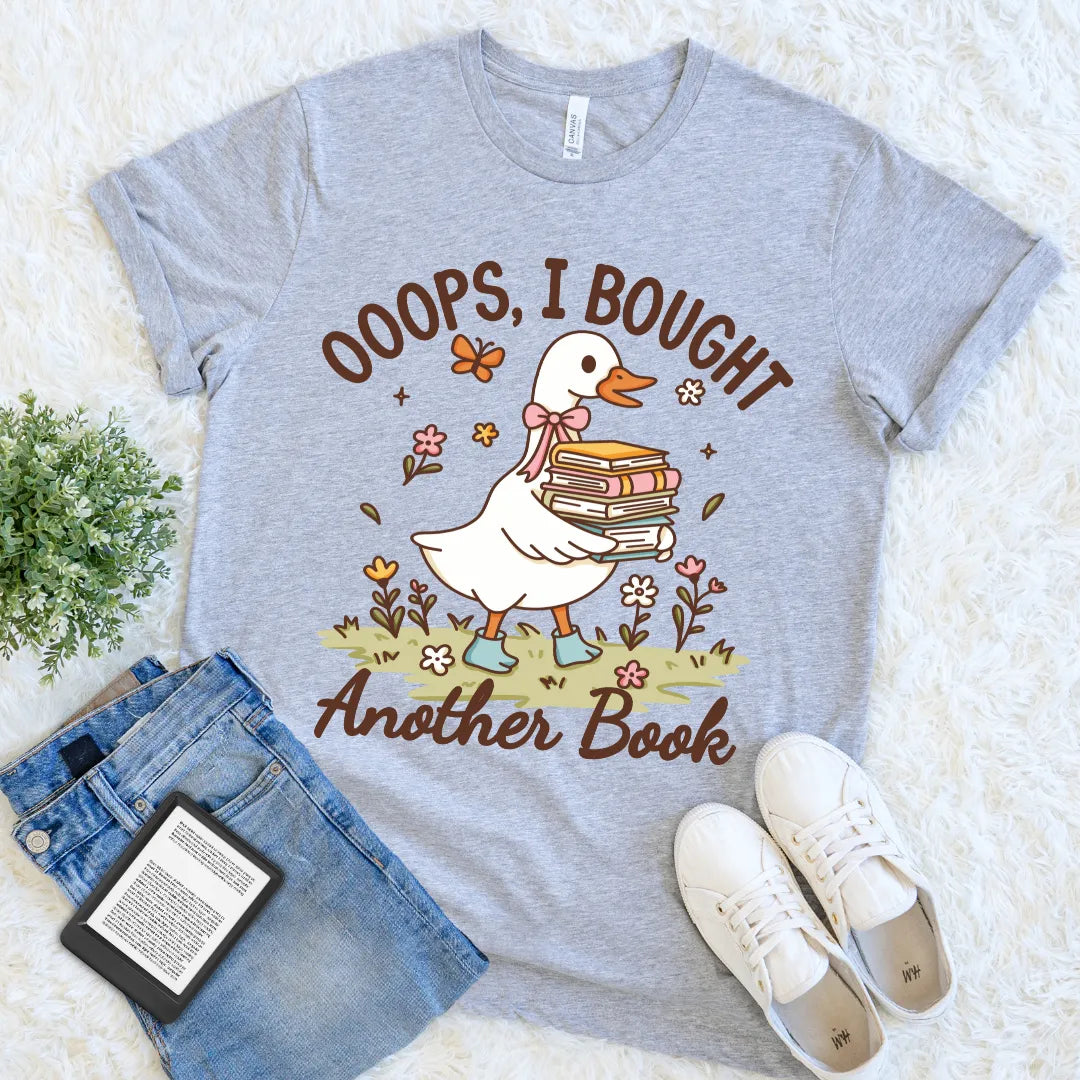 Gray t-shirt with a cartoon duck holding books and text 'Ooops, I bought another book' on a white surface with jeans and shoes.