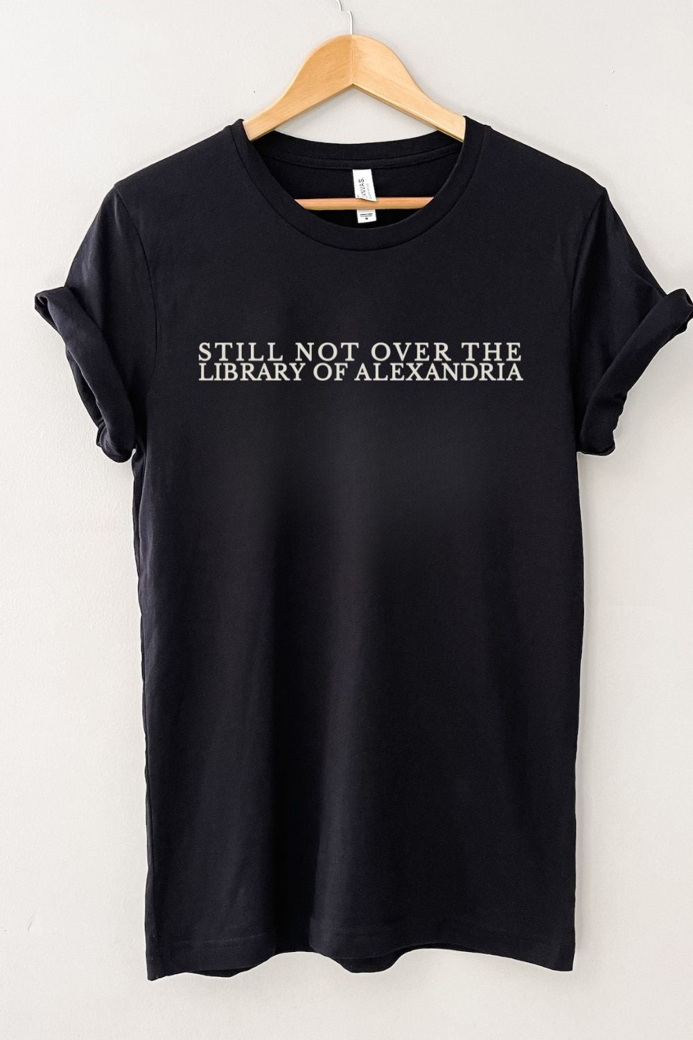 Black bookish t-shirt with 'Still Not Over the Library of Alexandria' text on a hanger against a white background