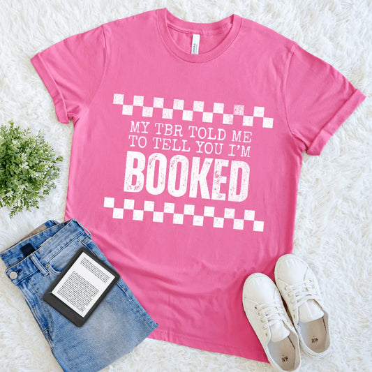 Flat lay of pink T-shirt with bold white text reading “My TBR told me to tell you I’m BOOKED”