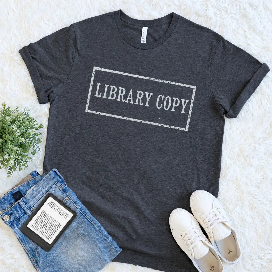 A dark heather gray book lover graphic t-shirt with 'LIBRARY COPY' stamp printed in distressed white text inside a rectangular border, styled in a flat lay with blue jeans, white sneakers, and a small green plant on a white faux fur background.