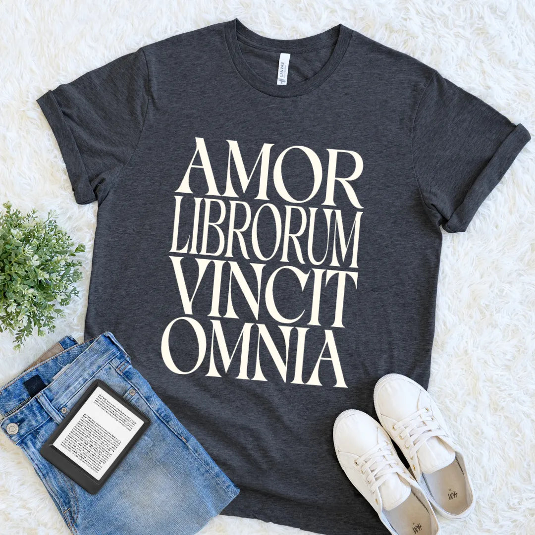 A dark charcoal gray graphic t-shirt with the phrase 'AMOR LIBRORUM VINCIT OMNIA' in classical Latin typography for readers and book lovers printed in large cream serif letters. The t-shirt is styled in a flat lay on a white faux fur background, accompanied by blue jeans, white sneakers, and a small green plant.