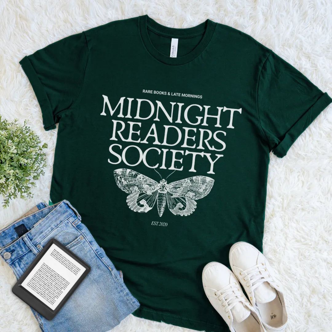 A forest green graphic t-shirt featuring the text 'MIDNIGHT READERS SOCIETY' and a detailed moth design, styled in a flat lay with blue jeans, a book, and white sneakers on a white faux fur background.