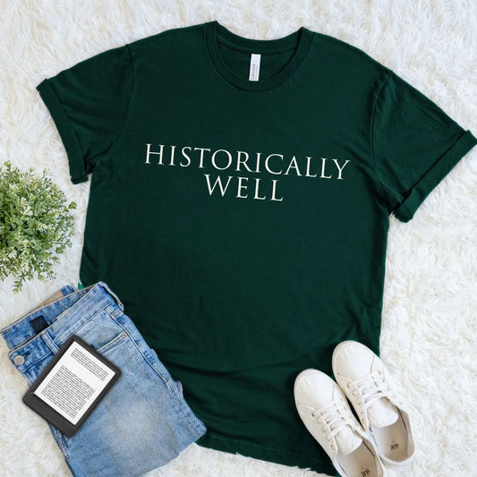 Forest green t-shirt with 'HISTORICALLY WELL' text on a light surface with jeans and sneakers.