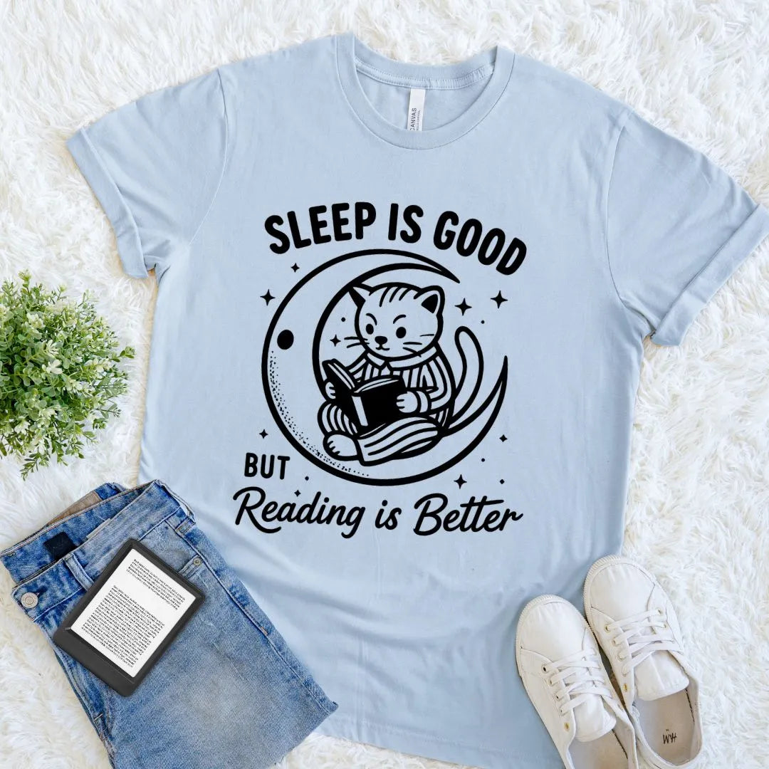 A light blue bookish graphic t-shirt laid flat on a white faux-fur surface, styled with folded blue jeans, a small green plant, and white sneakers. The shirt has a black graphic of a cat reading in a crescent moon with the text 'SLEEP IS GOOD BUT Reading is Better'.