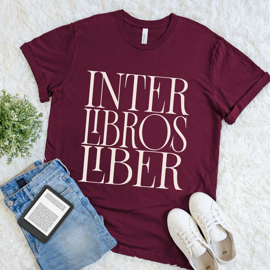 A maroon short-sleeve graphic T-shirt for book lovers laid flat on a white faux-fur surface, styled with blue jeans, a book, and white sneakers. The shirt has large cream text reading 'INTER LIBROS LIBER' across the front.