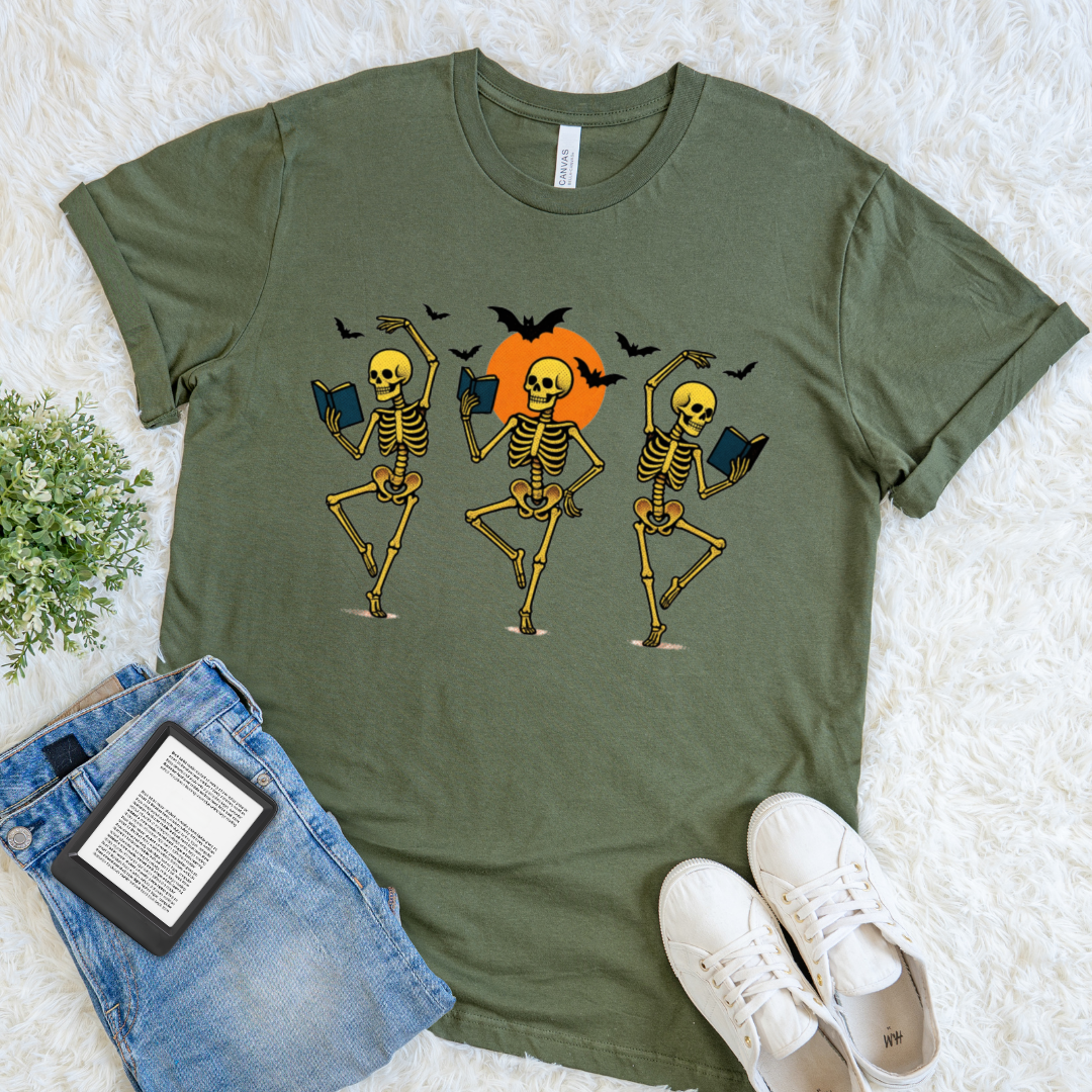 An olive green short-sleeve bookish t-shirt with a halloween graphic of three dancing skeletons in front of an orange sun and black bats, styled in a flat lay with blue jeans, a book, and white sneakers on a white faux fur background.