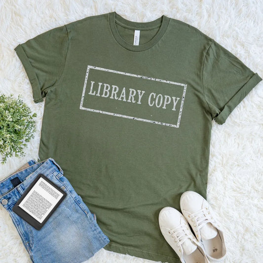An olive green graphic t-shirt with a distressed white 'LIBRARY COPY' stamp design, styled in a flat lay on a white faux fur rug with blue jeans, a book, and white sneakers.