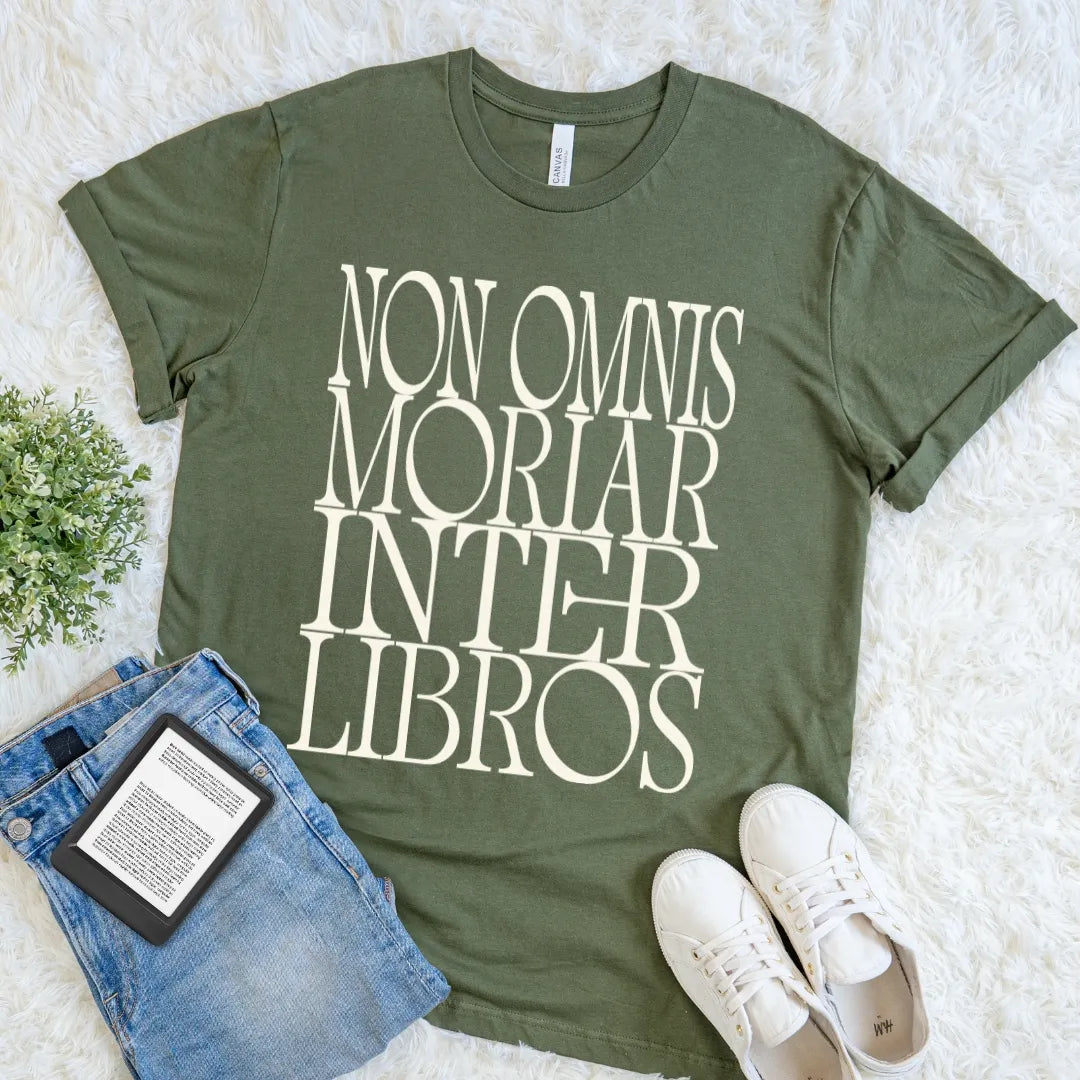 An olive green graphic t-shirt for devoted readers and bibliophiles with the text 'NON OMNIS MORIAR INTER LIBROS' in cream serif letters, styled in a flat lay on a white faux fur rug with blue jeans, a book, and white sneakers.