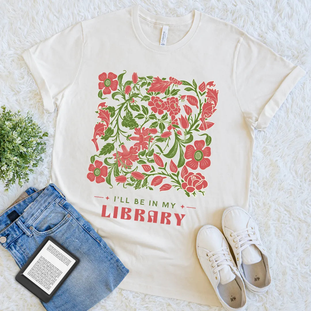 White t-shirt with floral design and 'I'll be in my library' text, placed on a textured surface with jeans, sneakers, and a book.