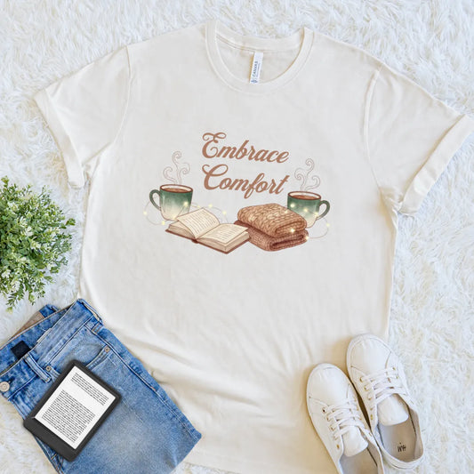 Soft cotton beige t-shirt with 'Embrace Comfort' graphic on a textured surface with jeans, sneakers, and a tablet, hygge-themed comfort shirt for readers