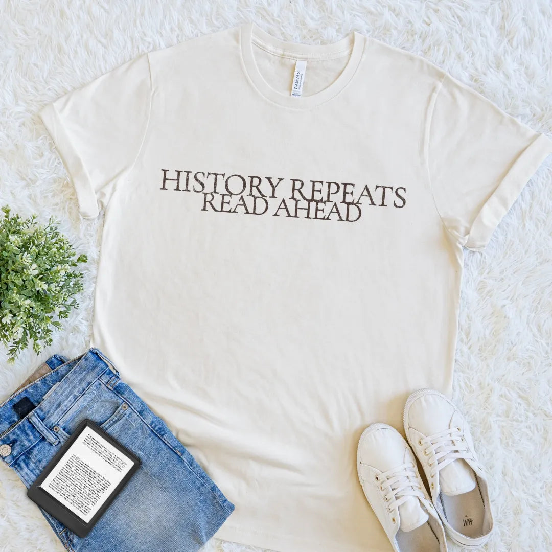 An off-white crewneck t-shirt with the text 'HISTORY REPEATS READ AHEAD' printed in dark brown serif letters, styled in a flat lay with blue jeans, white sneakers, a book, and a small green plant on a white shag rug.