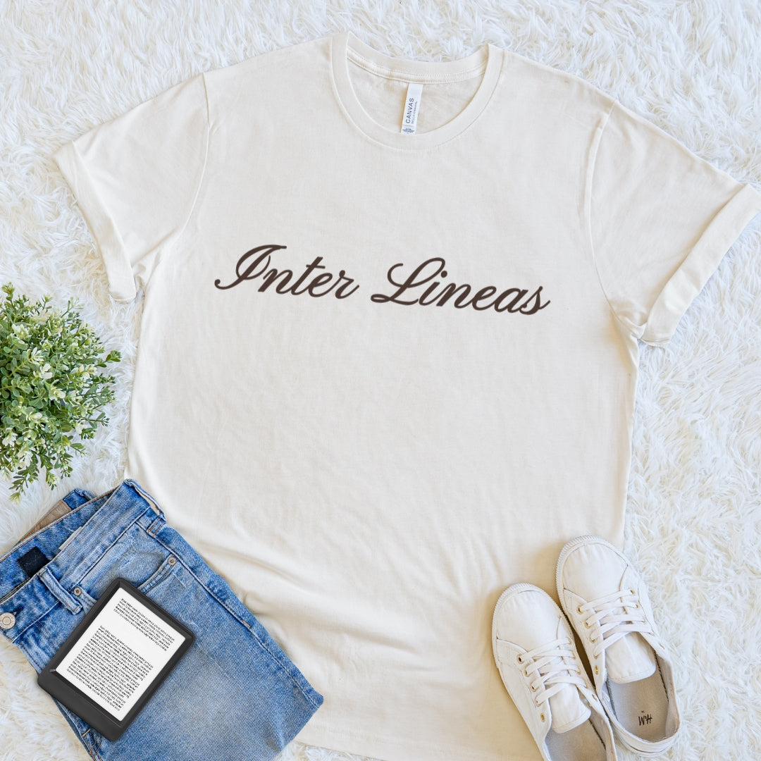 An off-white crewneck t-shirt with 'Inter Lineas' printed in dark brown cursive across the chest, styled in a flat lay with blue jeans, white sneakers, a book, and a small green plant on a white textured rug.