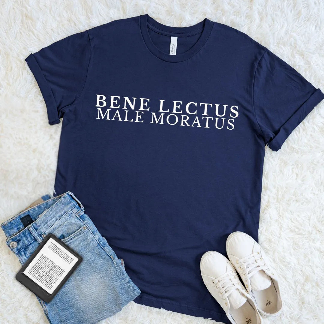 A navy blue graphic t-shirt a witty Latin reading quote in minimalist vintage typography Bene Lectus Male Moratus in white serif letters, styled in a flat lay on a white faux fur background with blue jeans, white sneakers, and a book.