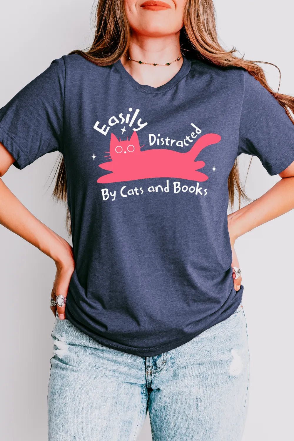 woman wearing a heather navy bookish t-shirt with pink cartoon cat and playful “easily distracted by cats and books” quote