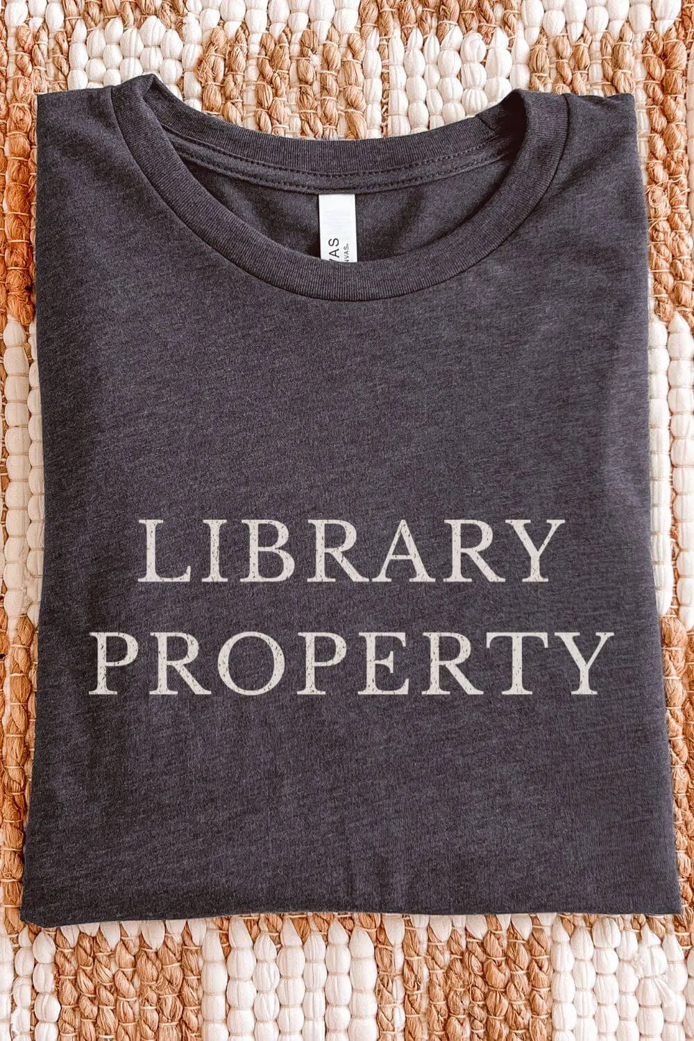 Folded dark gray heather t-shirt with the phrase “Library Property” printed in vintage typography, a minimalist book lover shirt styled on a woven background for a cozy library aesthetic.