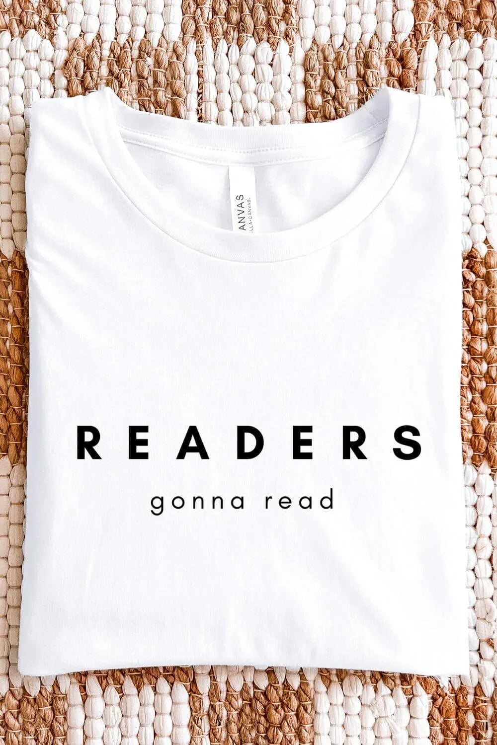 A white minimalist t-shirt with bold black text reading “Readers Gonna Read,” designed in a clean bookish aesthetic, folded on textile background