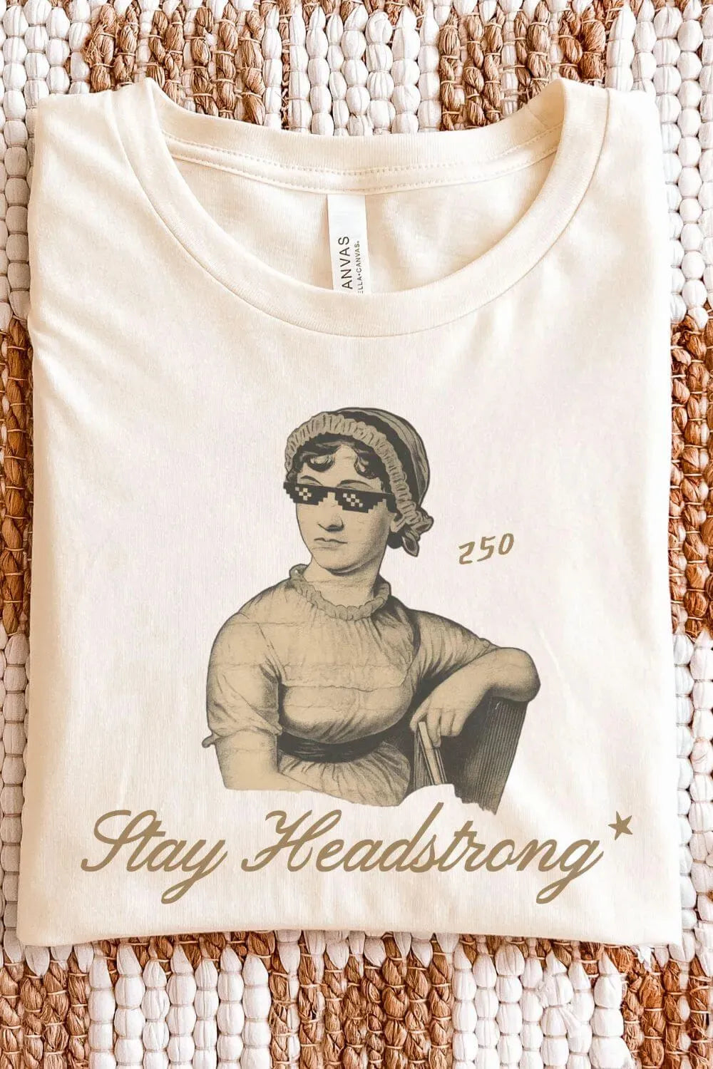Cream soft cotton bookish T-shirt with vintage Jane Austen wearing sunglasses illustration and “Stay Headstrong” quote, folded on textile surface