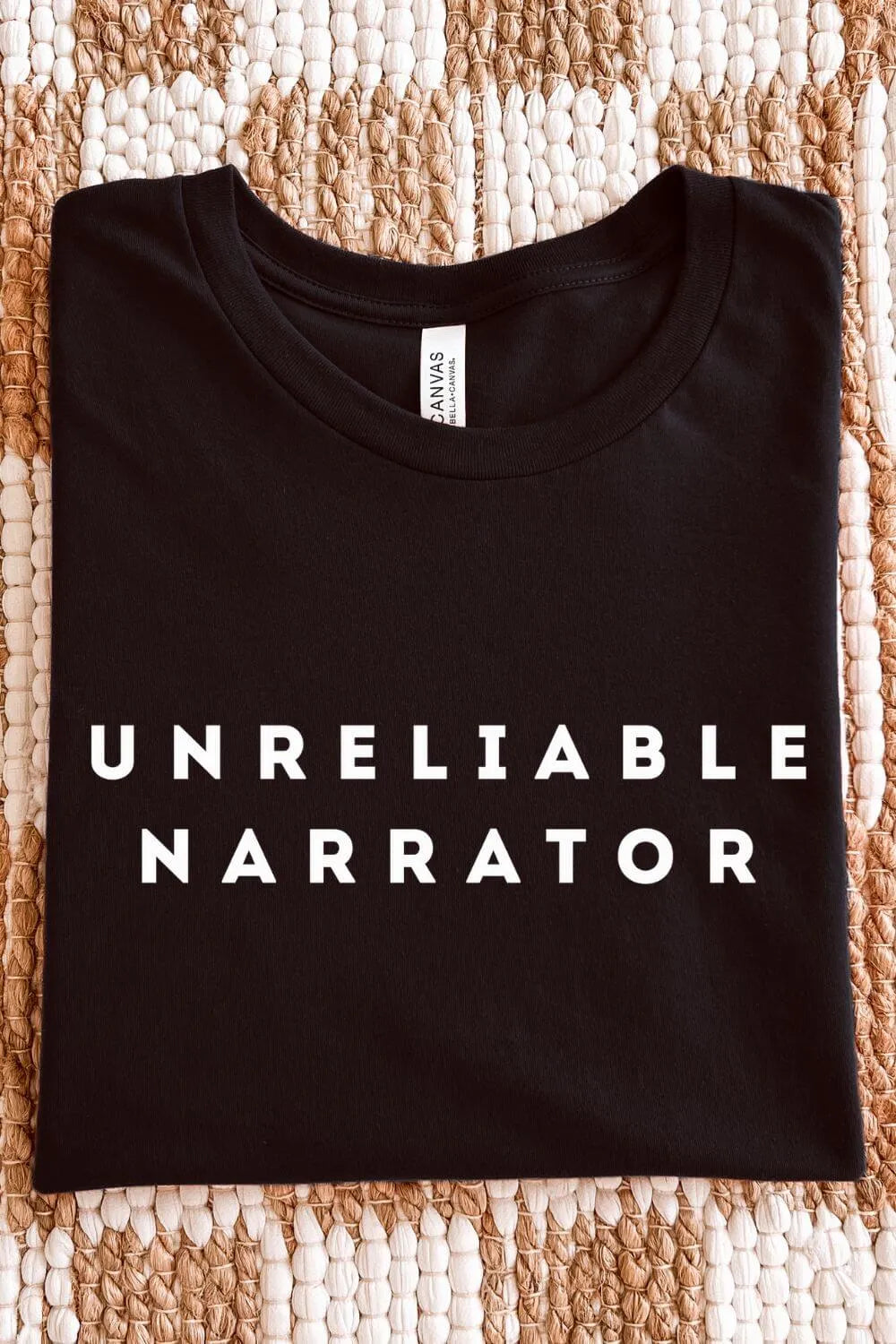 Black soft cotton t-shirt with 'UNRELIABLE NARRATOR' bookish humor text on a textured surface