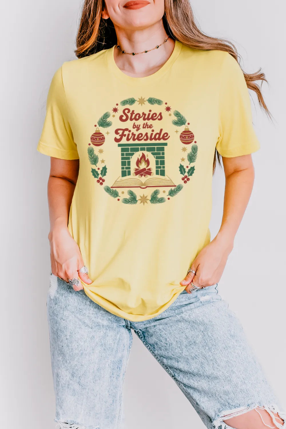Woman wearing yellow Christmas T-shirt  and jeans outfit with warm fireplace graphic with “Stories by the Fireside” text