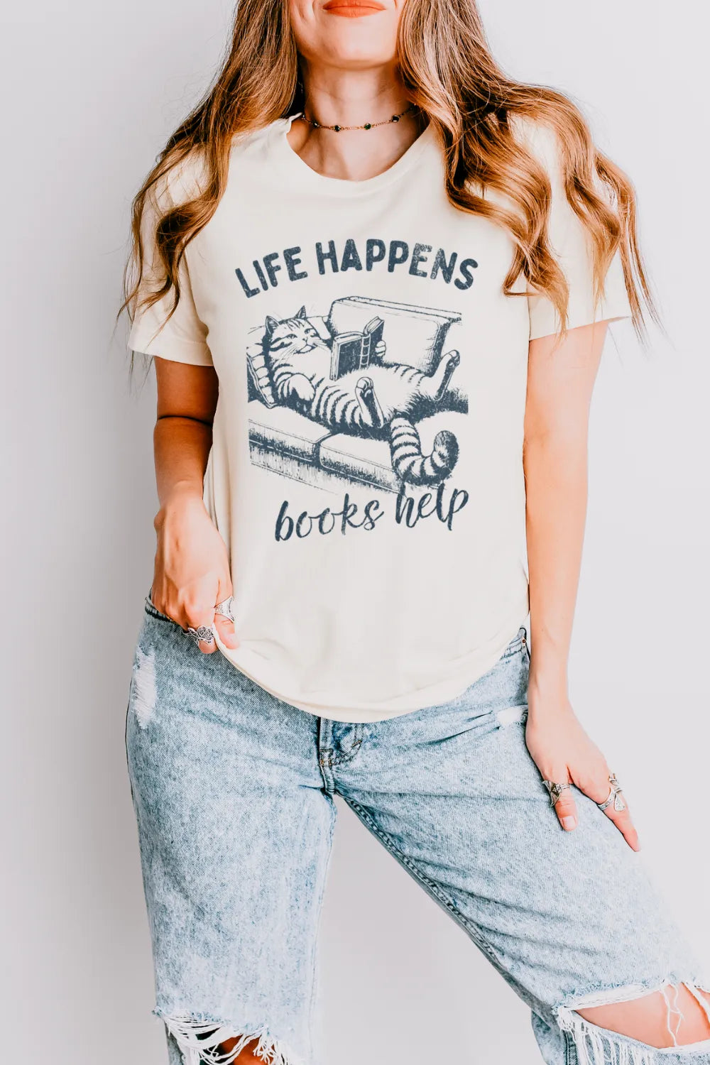 woman wearing cream T-shirt with cat reading on a couch illustration and “Life Happens Books Help” text