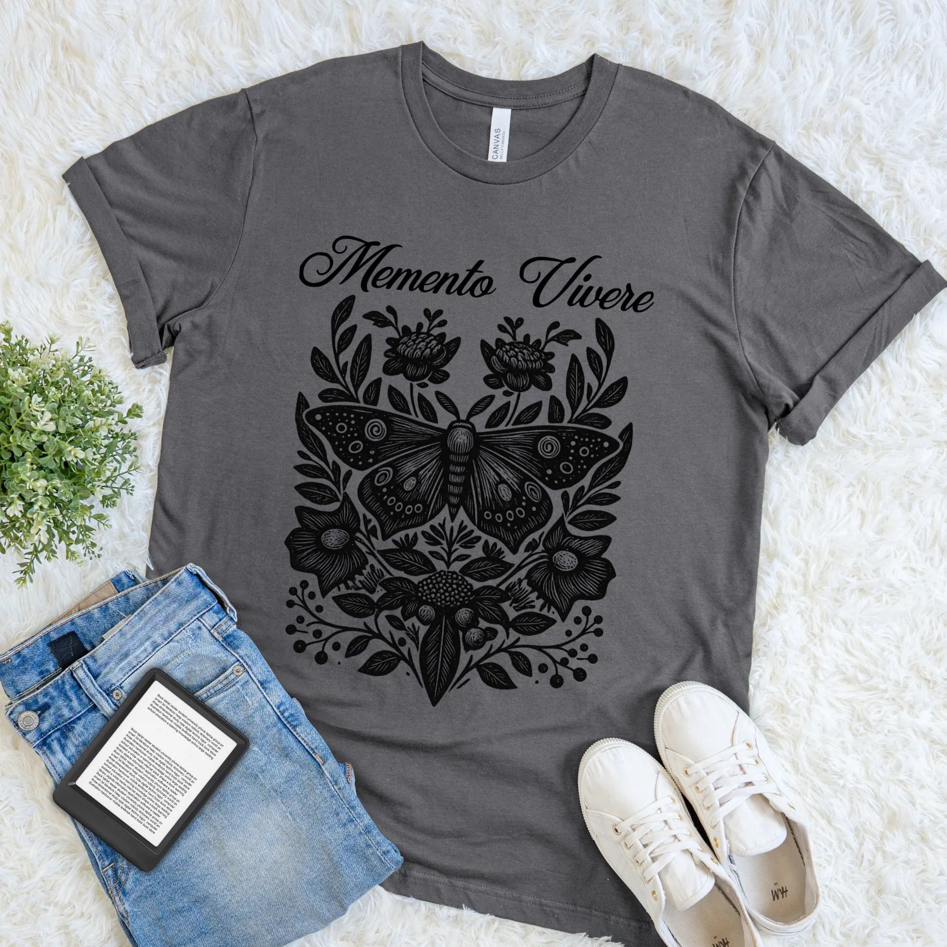 Gray t-shirt with black butterfly design and 'Memento Vivere' text, placed on a light surface with jeans and sneakers.