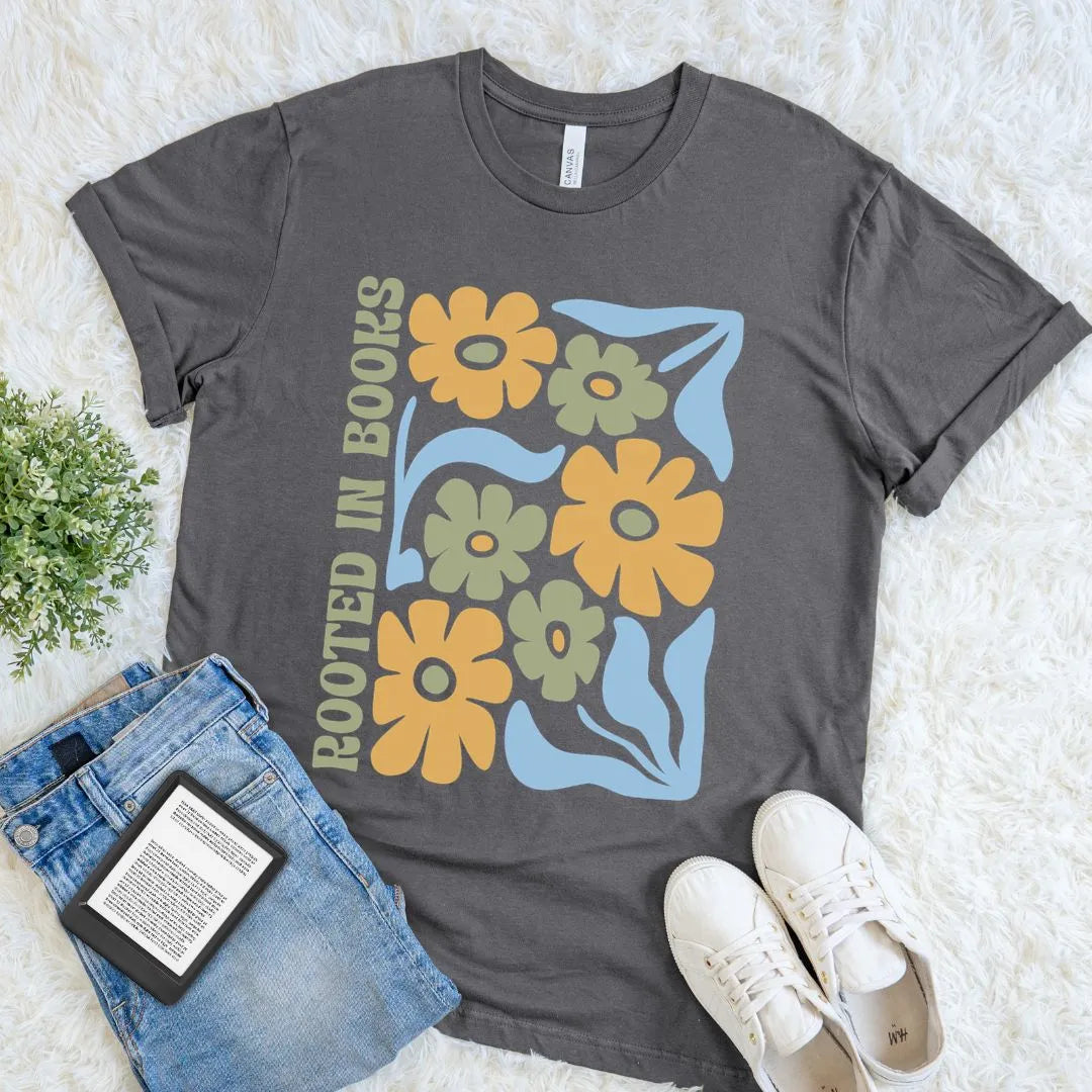 A asphalt grey  t-shirt featuring a retro floral design in mustard, sage, and light blue with the phrase ‘Rooted in Books’ along the side, styled flat with jeans and white sneakers