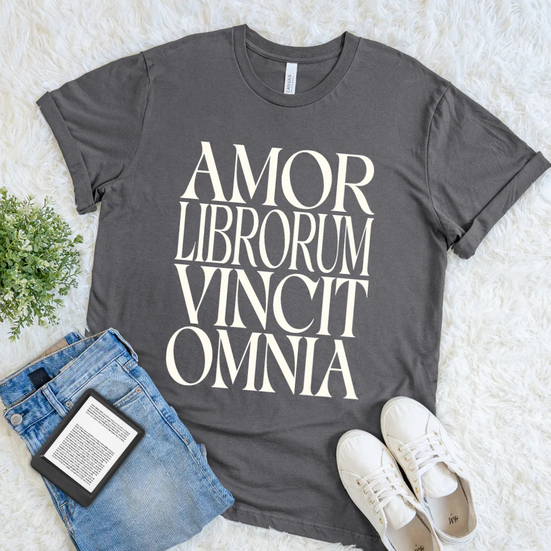 A dark gray book lover graphic t-shirt with the text 'AMOR LIBRORUM VINCIT OMNIA' in cream serif letters, styled in a flat lay with blue jeans, white sneakers, and a small green plant on a white faux fur background.