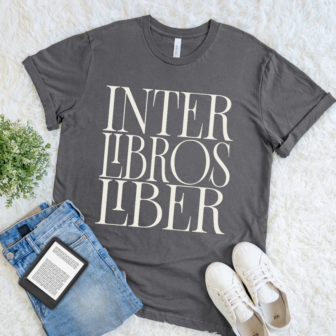 A dark gray graphic t-shirt with with bold Inter Libros Liber Latin phrase in elegant stacked cream serif lettering, styled in a flat lay on a white faux fur background with blue jeans, a book, and white sneakers.