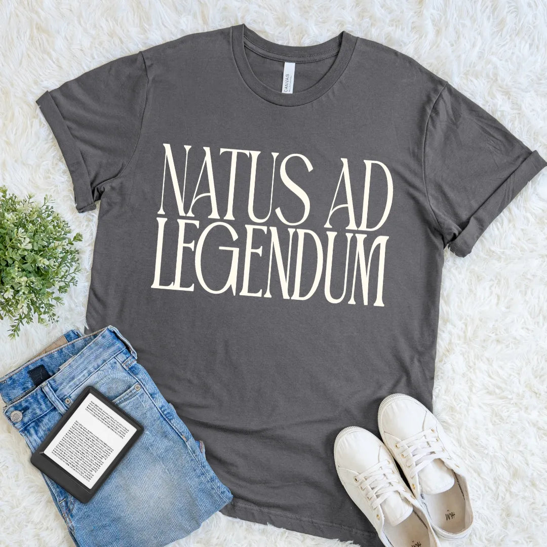  Charcoal literary T-shirt reader tee  featuring Natus ad Legendum in oversized cream serif typography, styled with jeans, an e-reader, and white sneakers on a soft rug.