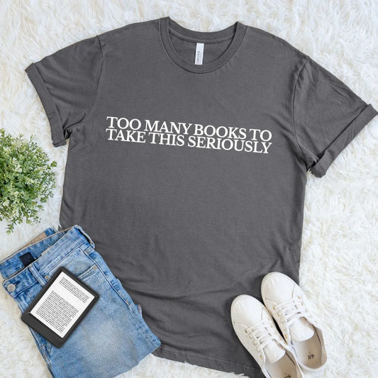 charcoal grey book lover T shirt featuring the quote Too Many Books to Take This Seriously styled with  jeans and white sneakers on white suface