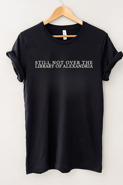 Black bookish t-shirt with 'Still Not Over the Library of Alexandria' text on a hanger against a white background
