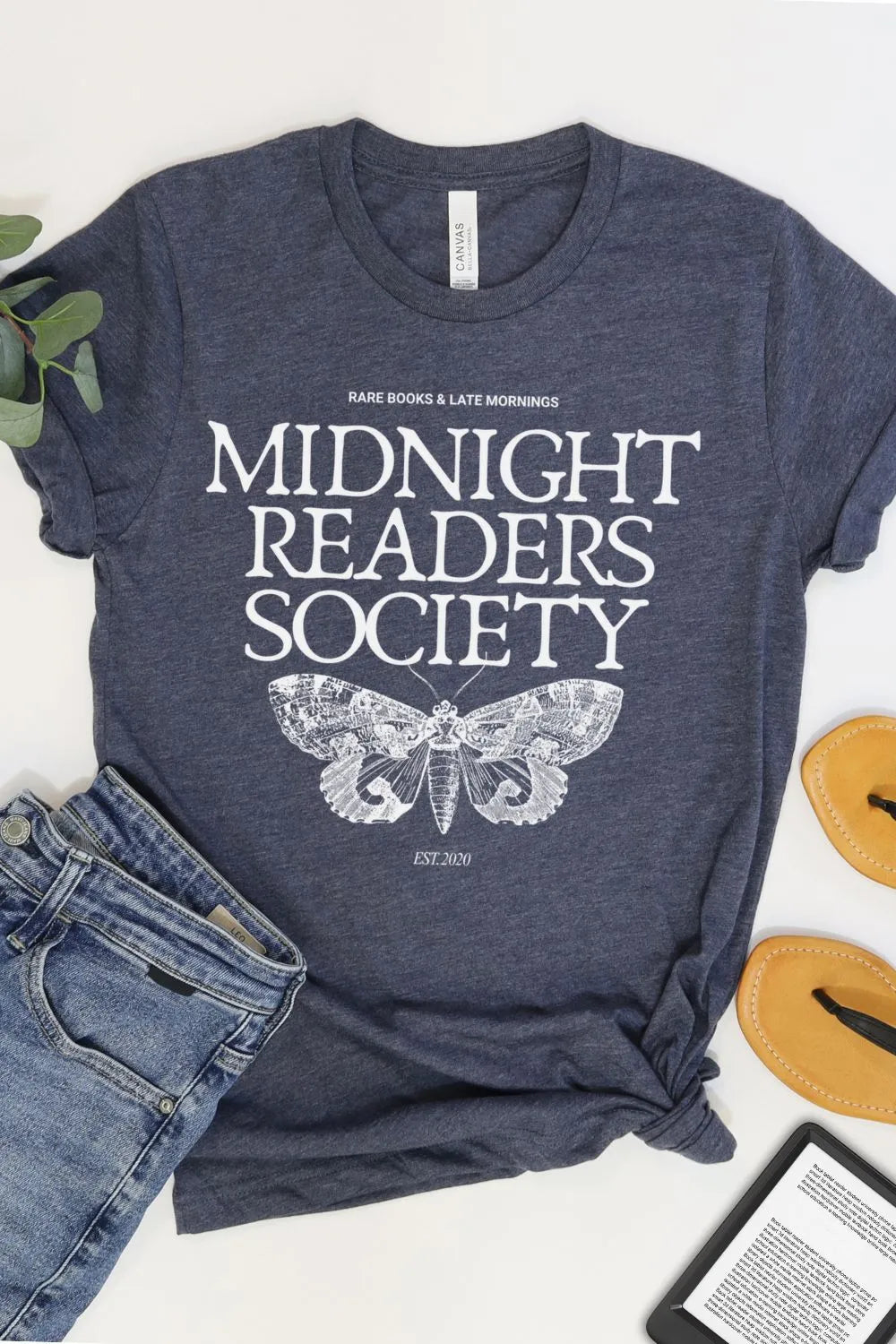 A soft heather navy bookish  t-shirt with vintage white text reading “Midnight Readers Society” and a detailed moth illustration, styled in a moody introvert aesthetic with jeans and ereader
