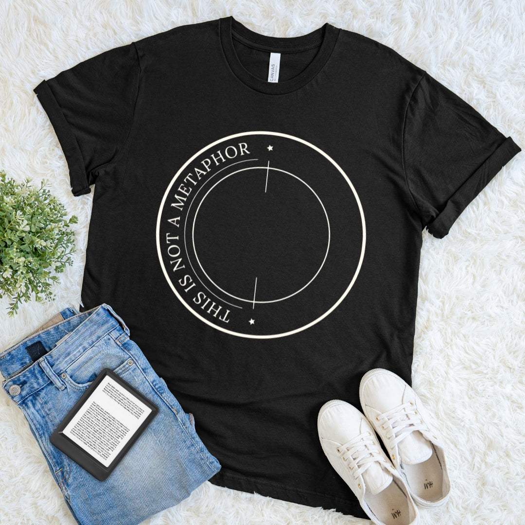 A black short-sleeve graphic T-shirt for readers laid flat on a white faux-fur surface, styled with blue jeans, white sneakers, and a small green plant. The shirt has a white circular line graphic with the text 'THIS IS NOT A METAPHOR' around the edge.