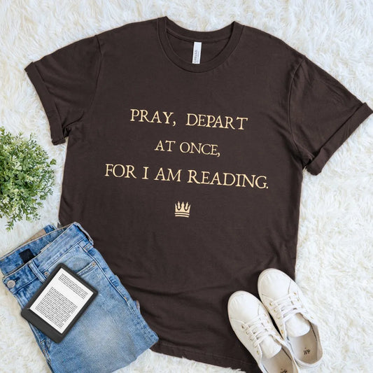 Brown t-shirt with text 'PRAY, DEPART AT ONCE, FOR I AM READING' and small crown illustration on a light background
