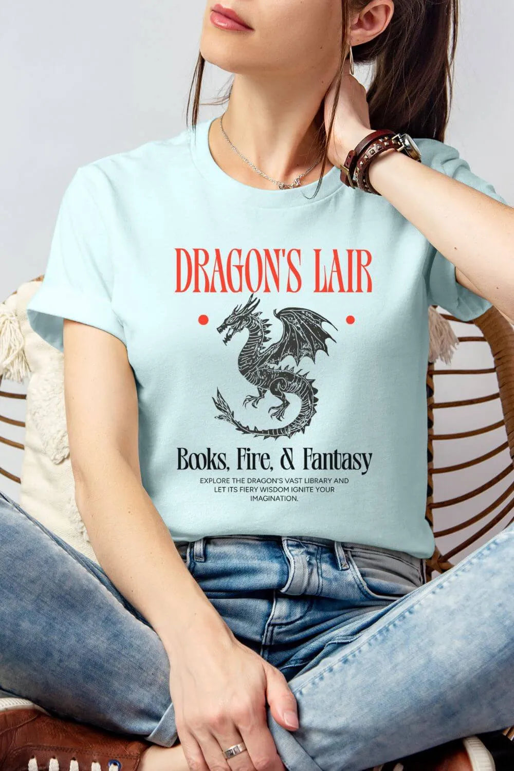 Ice blue book club graphic t-shirt with “Dragon’s Lair Books, Fire & Fantasy” text and dragon illustration, styled outfit with jeans wearing by model