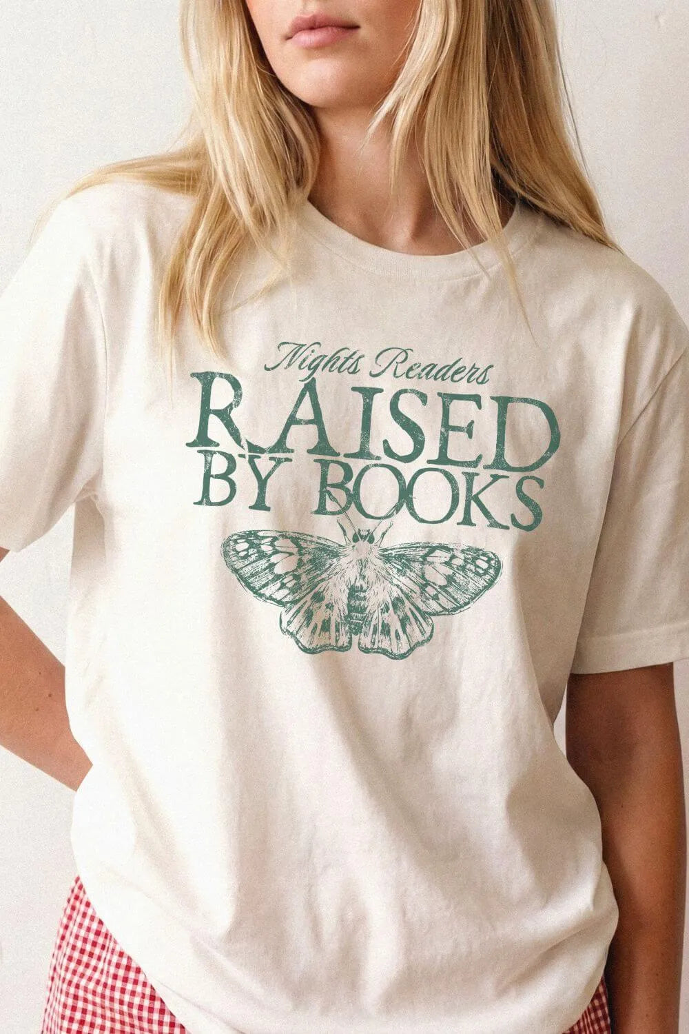 Woman wearing a beige bookish graphic t-shirt with “Raised By Books” typography and vintage moth illustration in soft academia aesthetic