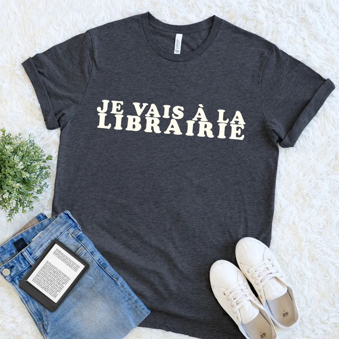 Dark grey heather literary T-shirt for Francophiles with Je Vais à La Librairie lettering, styled beside denim, an e-reader, white shoes, and greenery