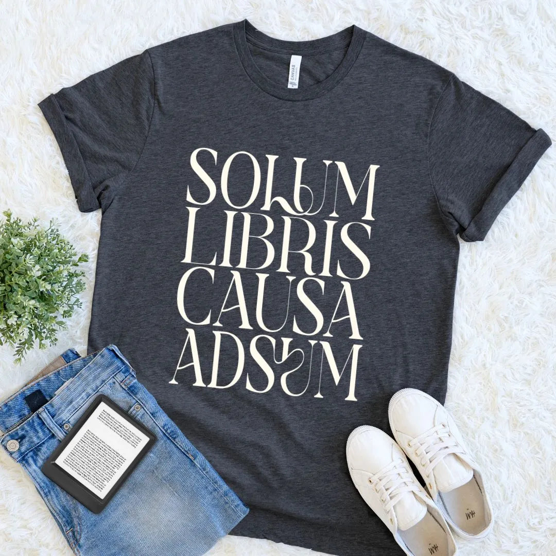A dark heather gray graphic T-shirt laid flat on a white faux-fur surface, styled with blue jeans, white sneakers, and a small green plant. The shirt has large white text reading 'SOLUM LIBRIS CAUSA ADS' across the front.