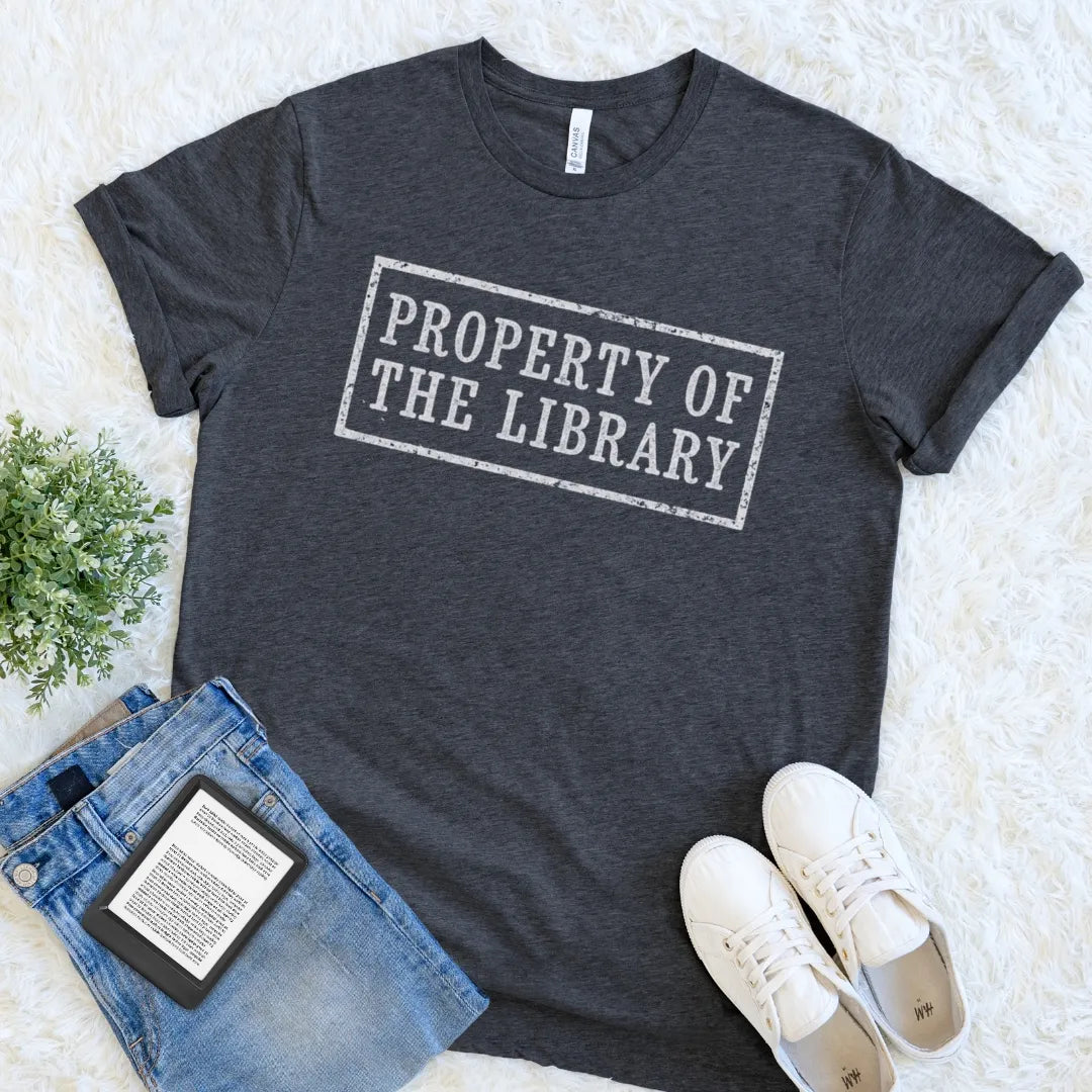 A dark heather gray bookish graphic t-shirt with the text 'PROPERTY OF THE LIBRARY' in a distressed white rectangular stamp design. The t-shirt is styled in a flat lay on a white faux fur background, accompanied by blue jeans, white sneakers, and a small green plant.