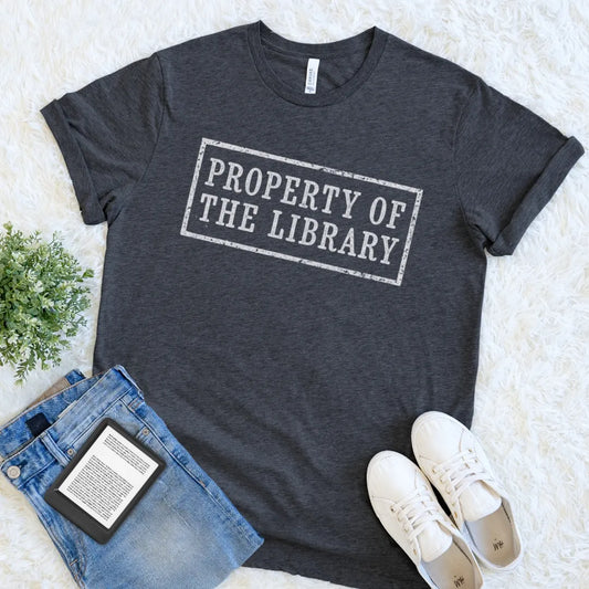 A dark heather gray bookish graphic t-shirt with the text 'PROPERTY OF THE LIBRARY' in a distressed white rectangular stamp design. The t-shirt is styled in a flat lay on a white faux fur background, accompanied by blue jeans, white sneakers, and a small green plant.