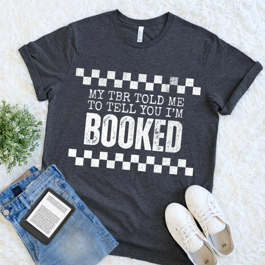 book lover shirt in dark grey heather with bold white text reading “My TBR told me to tell you I’m BOOKED” flat lay with denim and ereader for cozy reader outfit