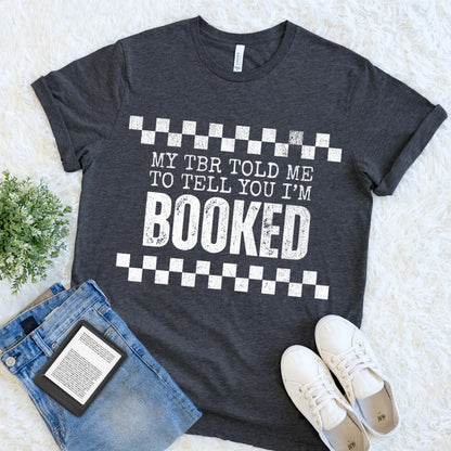 book lover shirt in dark grey heather with bold white text reading “My TBR told me to tell you I’m BOOKED” flat lay with denim and ereader for cozy reader outfit