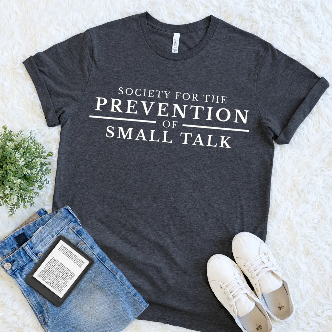 Dark gray t-shirt featuring the Society for the Prevention of Small Talk quote in cream lettering, paired with jeans and sneakers on a light background