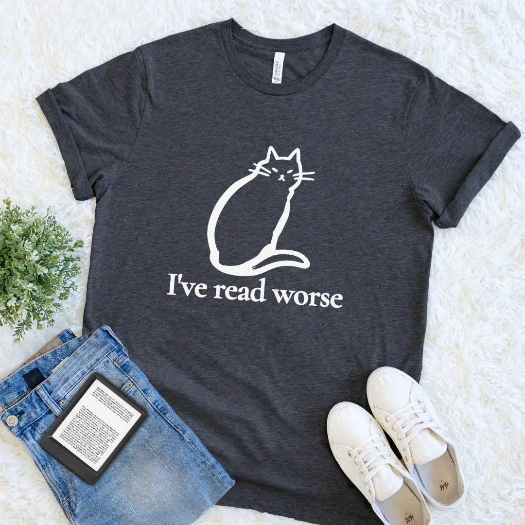 A dark heather gray bookish graphic t-shirt featuring a white line art cat and the humorous quote 'I've read worse'. The t-shirt is styled in a flat-lay arrangement on a white faux fur background, accompanied by blue jeans, a book, and white sneakers.