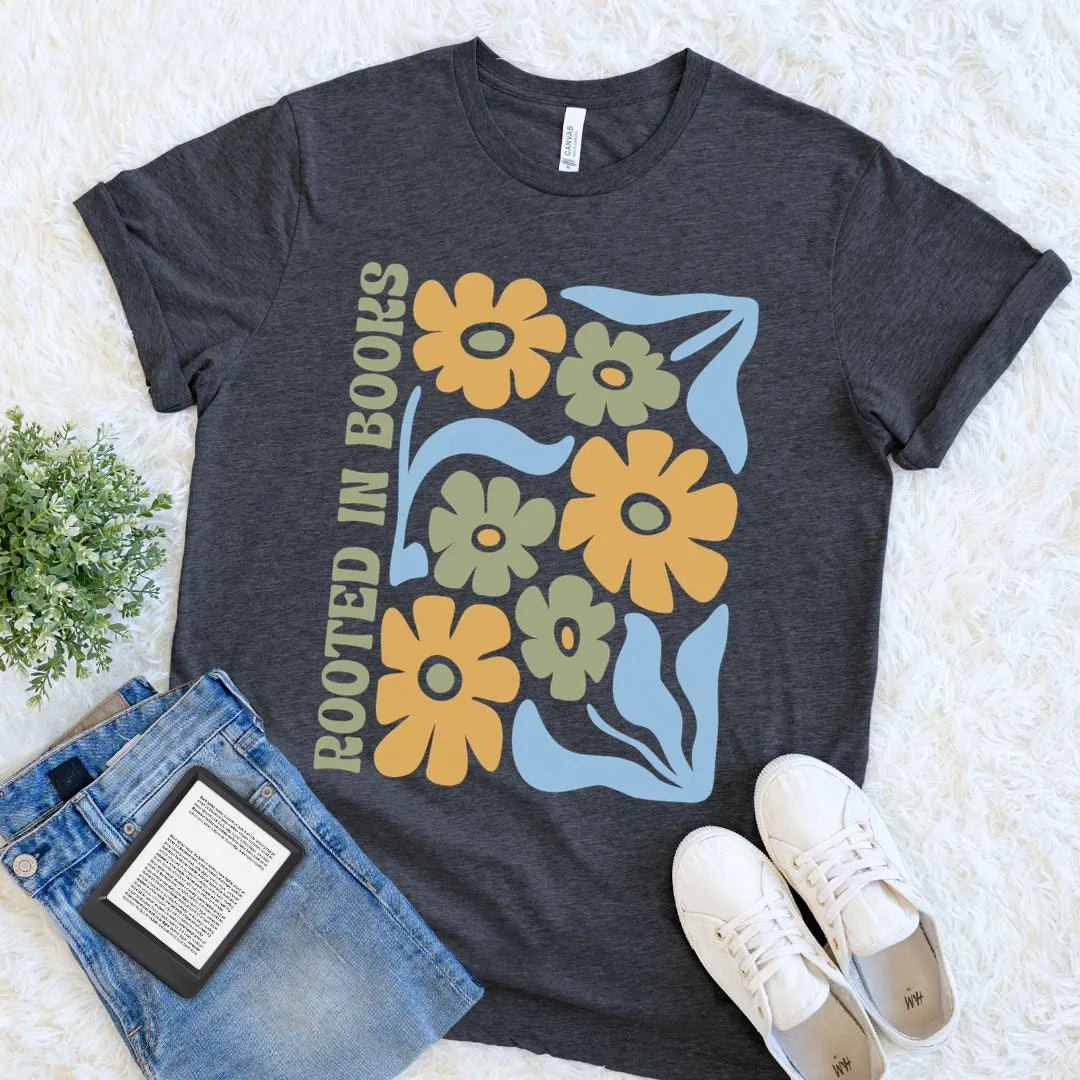 A  flat lay of dark grey heather  t-shirt featuring a boho  floral design in mustard, sage, and light blue with the ‘Rooted in Books’  text