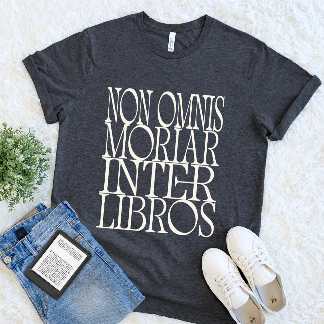 A dark heather gray graphic t-shirt with the phrase 'NON OMNIS MORIAR INTER LIBROS' printed in large cream serif letters. The t-shirt is styled in a flat lay on a white faux fur background, accompanied by blue jeans, white sneakers, and a small green plant.